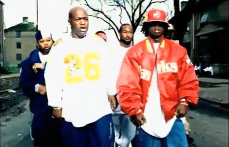 Lil Wayne with dark skin dreadlocks and a neutral expression positioned in the center of the frame. He is wearing a red and yellow Atlanta Hawks jacket with the word Hawks in large bold letters across the chest. He also wears a red and yellow baseball cap featuring the Atlanta Hawks logo. His right hand is raised in a gesture displaying a large ring.