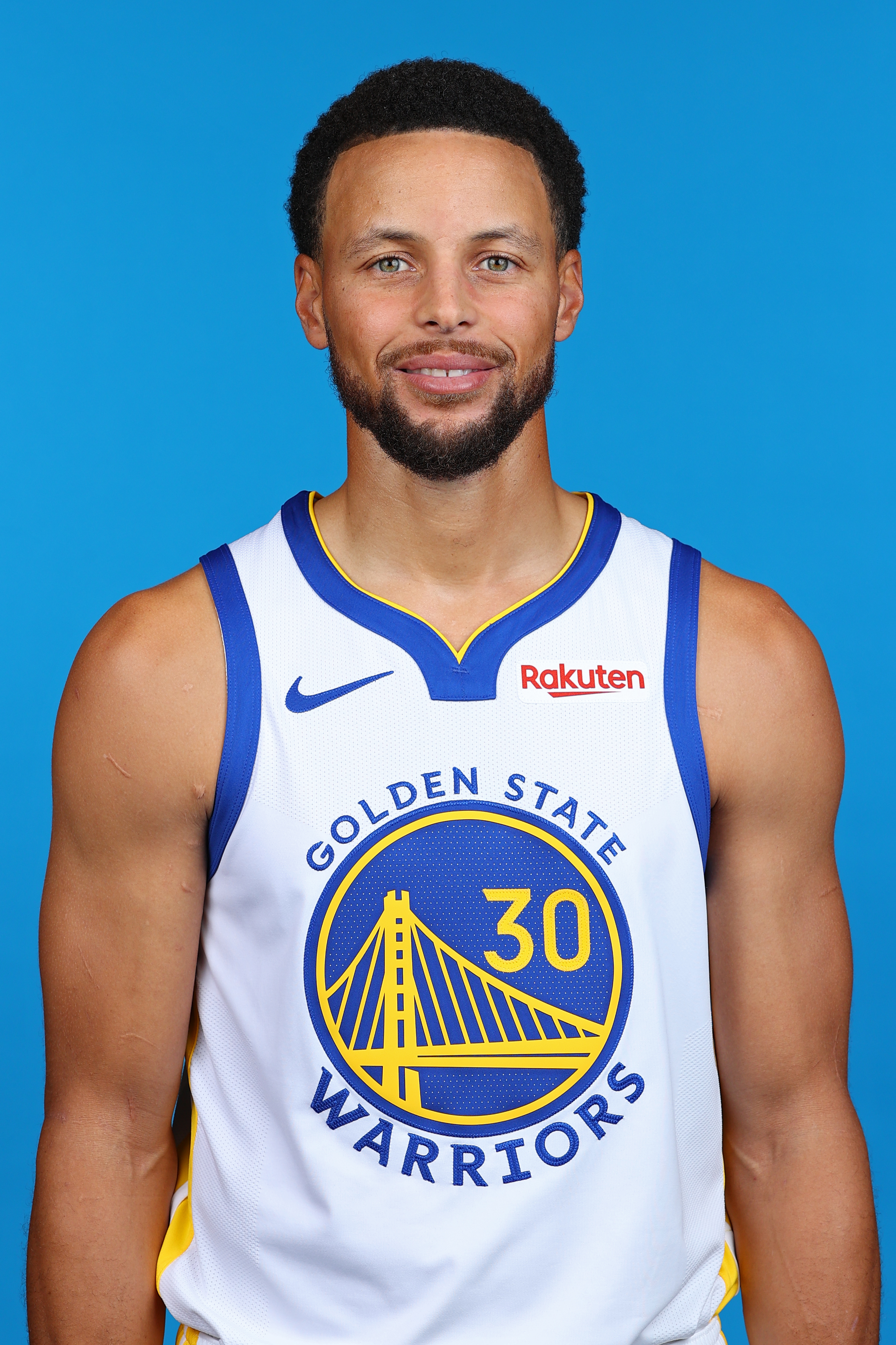 Professional NBA portrait photograph of Stephen Curry wearing Golden State Warriors #30 white home jersey with Nike and Rakuten sponsor logos. The subject is positioned against a solid blue background in a standard headshot pose. Curry displays a confident, slight smile with direct eye contact toward the camera. The image captures his upper body and shoulders, showcasing the distinctive Golden State Warriors uniform featuring the iconic Golden Gate Bridge logo in blue and gold. The lighting is even and professional, typical of official NBA team photography. The overall composition and quality suggest this is an official NBA/Warriors team photo used for roster documentation and media purposes.